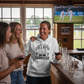 Three women in a pub watching cricket on TV, one wearing a Cows Corner hoodie with the funny cricket slogan 'Middle Order Collapse Survivor'. Cows Corner gifts are perfect for sport-mad fans, these gifts work brilliantly for birthdays, new baby celebrations, Father’s Day, Mother’s Day, Christmas, anniversaries, thank you gifts, end-of-season team awards, graduations, retirements, and just-because moments when you want to raise a smile. 
