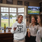 Three women in a pub setting, one wearing a hoodie with a unique design, with a TV showing a cricket match in the background. The lady is wearing a Cows Corner hoodie with the funny cricket slogan 'Bowled Over by Love'. Cows Corner gifts are perfect for sport-mad fans, these gifts work brilliantly for birthdays, new baby celebrations, Father’s Day, Mother’s Day, Christmas, anniversaries, thank you gifts, end-of-season team awards, graduations, retirements, and just-because moments 
