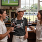 Three women in cricket attire are enjoying drinks in a pub with a TV showing a cricket match.  The lady is wearing a Cows Corner t-shirt with the funny cricket saying on the front which says 'Bowled & Bamboozled And Still Looking Better Than You'