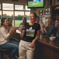 Woman in a sports bar holding a glass of wine, surrounded by friends watching a game on TV with the lady wearing a Cows corner t-shirt with the slogan on the front saying 'Bowled & Bamboozled and still looking better than you'.  Cows Corner gifts are perfect for sport-mad fans who love a bit of humour and personality in their presents. They’re ideal for friends, partners, parents, coaches, teammates, and proud mums and dads who live and breathe sport, whether they play, watch from the sidelines