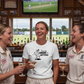 Three women in cricket uniforms enjoying drinks in a bar with a cricket match on TV.  The lady is wearing a Cows Corner T-shirt with the funny cricket slogan on the front that reads 'I Bat in the Middle of a Hat-trick'.  Cows Corner gifts are perfect for sport-mad fans, these gifts work brilliantly for birthdays, new baby celebrations, Father’s Day, Mother’s Day, Christmas, anniversaries, thank you gifts, end-of-season team awards, graduations, retirements, and just-because moments.