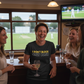 People in a pub watching cricket on TV, one person holding a glass of wine with a humorous t-shirt.  The lady is wearing a Cows Corner t-shirt with the funny cricket slogan 'I Don't Block, Only Bash' on the front.