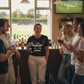 Group of people in a pub watching cricket on TV, one person holding a glass with a humorous t-shirt with the Cows Corner funny slogan on the front saying 'I play better with a stiff handle'.