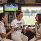 Three women in cricket attire sitting together, smiling and holding drinks in a sports bar.  The lady is wearing a Cows Corner T-shirt with the funny cricket slogan on the front that reads 'Legends Are Made in the Nets'. Cows Corner gifts are perfect for sport-mad fans, these gifts work brilliantly for birthdays, new baby celebrations, Father’s Day, Mother’s Day, Christmas, anniversaries, thank you gifts, end-of-season team awards, graduations, retirements, and just-because moments 
