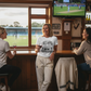 Three women in a pub watching cricket on television, one wearing a 'Collaps Survivor' t-shirt.  She is wearing a Cows Corner T-shirt with the funny cricket slogan on the front that reads 'Middle-Order Collapse Survivor'.  Cows Corner gifts are perfect for sport-mad fans, these gifts work brilliantly for birthdays, new baby celebrations, Father’s Day, Mother’s Day, Christmas, anniversaries, thank you gifts, end-of-season team awards, graduations, retirements, and just-because moments.