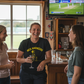 Three women in a casual setting with a TV showing a sports game in the background.  The lady is wearing a Cows Corner t-shirt with the funny cricket slogan on front saying 'Nice Bouncers'. 