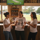 Three women in a pub watching a cricket match on TV, holding drinks. The lady is wearing a Cows Corner T-shirt with the cricket coach slogan on the front that reads 'To My Coach Who Made Me Believe in My First Cover Drive – You’re a Legend'. Cows Corner gifts are perfect for sport-mad fans, these gifts work brilliantly for birthdays, new baby celebrations, Father’s Day, Mother’s Day, Christmas, anniversaries, thank you gifts, end-of-season team awards, graduations, retirements, and just-because moments