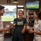 People in sports attire enjoying drinks in a bar with a cricket match on TV.  The lady is wearing a Cows Corner t-shirt with the funny cricket saying on the front saying 'Wanna See My Googly?  Funny Cricket T-Shirt'