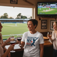 People in a bar watching a sports game on TV with one person wearing a unique t-shirt.  The lady is wearing a Cows Corner t-shirt with the cricket coach slogan on the front that reads 'You Built My Technique My Confidence and My Game – Thank You'. Cows Corner gifts are perfect for sport-mad fans, these gifts work brilliantly for birthdays, new baby celebrations, Father’s Day, Mother’s Day, Christmas, anniversaries, thank you gifts, end-of-season team awards, graduations, retirements, and just-because moment