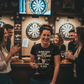 Group of people in a bar with dartboards and a woman wearing a t-shirt with a humorous darts player quote. The lady is wearing a Cows Corner t-shirt with the funny darts slogan 'Behind Every Great Darts Player Is An Empty Pint Glass'. Cows Corner gifts are perfect for sport-mad fans, these gifts work brilliantly for birthdays, new baby celebrations, Father’s Day, Mother’s Day, Christmas, anniversaries, thank you gifts, end-of-season team awards, graduations, retirements, and just-because moments 