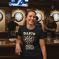 Woman wearing a black t-shirt with a dartboard design and text, standing in a bar setting. The lady is wearing a Cows Corner t-shirt with the funny darts slogan 'Darts Because Therapy is Expensive'.  Cows Corner gifts are perfect for sport-mad fans, these gifts work brilliantly for birthdays, new baby celebrations, Father’s Day, Mother’s Day, Christmas, anniversaries, thank you gifts, end-of-season team awards, graduations, retirements, and just-because moments when you want to raise a smile. 
