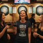 Three women in a pub setting with dartboards and a TV in the background. The lady is wearing a Cows Corner t-shirt with the funny darts slogan 'I Bring the Darts You Bring the Excuses'. Cows Corner gifts are perfect for sport-mad fans, these gifts work brilliantly for birthdays, new baby celebrations, Father’s Day, Mother’s Day, Christmas, anniversaries, thank you gifts, end-of-season team awards, graduations, retirements, and just-because moments when you want to raise a smile. 
