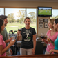 Group of people in a golf-themed bar with a woman in a humorous t-shirt holding a glass of wine. The lady is wearing a Cows Corner t-shirt with the funny golf slogan 'I Got A Hole-In-One… My Mates Got Trauma'. Cows Corner gifts are perfect for sport-mad fans, these gifts work brilliantly for birthdays, new baby celebrations, Father’s Day, Mother’s Day, Christmas, anniversaries, thank you gifts, end-of-season team awards, graduations, retirements, and just-because moments when you want to raise a smile. 
