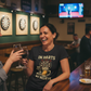Woman in a bar wearing a t-shirt with a dartboard design, holding a glass of wine. The lady is wearing a Cows Corner t-shirt with the funny darts slogan 'In Darts We Trust, in Beer We Believe'. Cows Corner gifts are perfect for sport-mad fans, these gifts work brilliantly for birthdays, new baby celebrations, Father’s Day, Mother’s Day, Christmas, anniversaries, thank you gifts, end-of-season team awards, graduations, retirements, and just-because moments when you want to raise a smile. 
