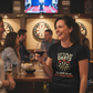 Woman in a bar wearing a 'Let's Play Darts' shirt, holding a glass of wine. The lady is wearing a Cows Corner t-shirt with a funny darts slogan 'Lets Play Darts I'll Show You My Best Shaft Work'. Cows Corner gifts are perfect for sport-mad fans, these gifts work brilliantly for birthdays, new baby celebrations, Father’s Day, Mother’s Day, Christmas, anniversaries, thank you gifts, end-of-season team awards, graduations, retirements, and just-because moments when you want to raise a smile. 

