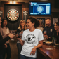 Woman in a bar wearing a white t-shirt with a logo, holding a glass of wine, surrounded by friends. The lady is wearing a Cows Corner t-shirt with the funny darts slogan 'One Hand For The Pint, One Hand For The Darts'. Cows Corner gifts are perfect for sport-mad fans, these gifts work brilliantly for birthdays, new baby celebrations, Father’s Day, Mother’s Day, Christmas, anniversaries, thank you gifts, end-of-season team awards, graduations, retirements, and just-because moments when you want to raise a sm