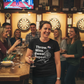 Woman in a bar wearing a t-shirt with text, surrounded by friends and dartboards. The lady is wearing a Cows Corner t-shirt with a funny darts slogan 'Throw. Miss. Swear. Repeat'. Cows Corner gifts are perfect for sport-mad fans, these gifts work brilliantly for birthdays, new baby celebrations, Father’s Day, Mother’s Day, Christmas, anniversaries, thank you gifts, end-of-season team awards, graduations, retirements, and just-because moments when you want to raise a smile. 
