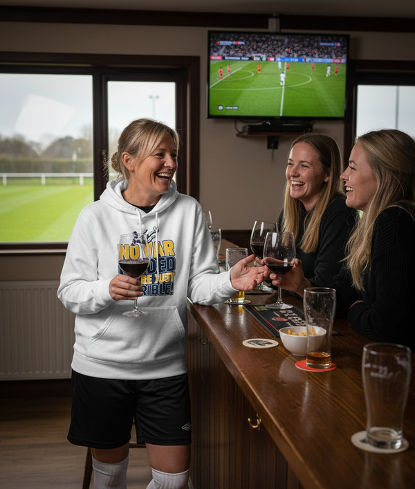 Three women in a bar watching a sports match on TV, holding drinks. The lady was wearing a Cows Corner hoodie with the funny football slogan 'No VAR Needed You’re Just Terrible'. Cows Corner gifts are perfect for sport-mad fans, these gifts work brilliantly for birthdays, new baby celebrations, Father’s Day, Mother’s Day, Christmas, anniversaries, thank you gifts, end-of-season team awards, graduations, retirements, and just-because moments when you want to raise a smile. 
