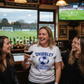 Woman in a sports bar with friends, holding a glass of red wine, wearing a t-shirt with a soccer-themed message.  The lady is wearing a Cows Corner T-shirt with the funny football slogan on the front that reads 'Dribble You Can Barely Walk Straight!'.  Cows Corner gifts are perfect for sport-mad fans, these gifts work brilliantly for birthdays, new baby celebrations, Father’s Day, Mother’s Day, Christmas, anniversaries, thank you gifts, end-of-season team awards, graduations, retirements, and just-because