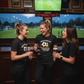 Three women in a bar watching sports on TV with drinks.  The lady is wearing a Cows Corner t-shirt with the football slogan on the front that reads 'For Believing in Me When I Was Still Chasing Cones – Thank You, Coach'. Cows Corner gifts are perfect for sport-mad fans, these gifts work brilliantly for birthdays, new baby celebrations, Father’s Day, Mother’s Day, Christmas, anniversaries, thank you gifts, end-of-season team awards, graduations, retirements, and just-because moments 