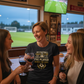 Three women in a sports bar watching a football match on television, holding wine glasses.  The lady is wearing a Cows Corner t-shirt with the football slogan on the front that reads 'My Football Coach Is the Best in the World, I’d Know I’ve Played Under the Rest'. Cows Corner gifts are perfect for sport-mad fans, these gifts work brilliantly for birthdays, new baby celebrations, Father’s Day, Mother’s Day, Christmas, anniversaries, thank you gifts, end-of-season team awards, graduations, retirements 