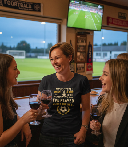 Three women in a sports bar watching a football match on television, holding wine glasses.  The lady is wearing a Cows Corner t-shirt with the football slogan on the front that reads 'My Football Coach Is the Best in the World, I’d Know I’ve Played Under the Rest'. Cows Corner gifts are perfect for sport-mad fans, these gifts work brilliantly for birthdays, new baby celebrations, Father’s Day, Mother’s Day, Christmas, anniversaries, thank you gifts, end-of-season team awards, graduations, retirements 