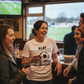 People in a pub watching a sports match, one person wearing a Cows Corner T-shirt with the funny football slogan on the front that reads 'Play Football Not Fortnite'.  Cows Corner gifts are perfect for sport-mad fans, these gifts work brilliantly for birthdays, new baby celebrations, Father’s Day, Mother’s Day, Christmas, anniversaries, thank you gifts, end-of-season team awards, graduations, retirements, and just-because moments when you want to raise a smile.  
