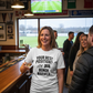 Woman wearing a t-shirt with a humorous sports-related message in a pub setting.  The lady is wearing a Cows Corner t-shirt with the funny football slogan on the front that reads 'Your Best Position? Bench Warmer'. Cows Corner gifts are perfect for sport-mad fans, these gifts work brilliantly for birthdays, new baby celebrations, Father’s Day, Mother’s Day, Christmas, anniversaries, thank you gifts, end-of-season team awards, graduations, retirements, and just-because moments when you want to raise a smile.