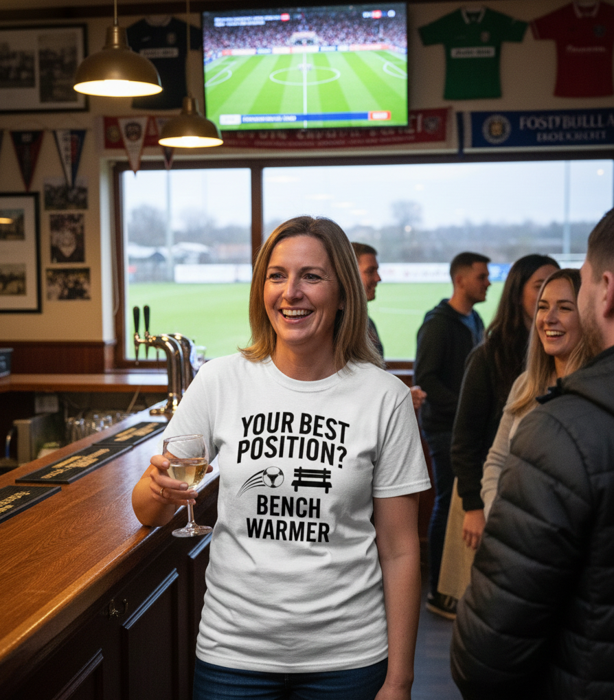 Woman wearing a t-shirt with a humorous sports-related message in a pub setting.  The lady is wearing a Cows Corner t-shirt with the funny football slogan on the front that reads 'Your Best Position? Bench Warmer'. Cows Corner gifts are perfect for sport-mad fans, these gifts work brilliantly for birthdays, new baby celebrations, Father’s Day, Mother’s Day, Christmas, anniversaries, thank you gifts, end-of-season team awards, graduations, retirements, and just-because moments when you want to raise a smile.