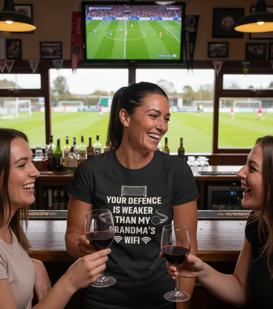 Three women in a bar watching a sports game on TV, one wearing a humorous t-shirt.  The lady is wearing a Cows Corner T-shirt with the funny football slogan on the front that reads 'Your Defence Is Weaker Than My Grandma’s Wifi'.  Cows Corner gifts are perfect for sport-mad fans, these gifts work brilliantly for birthdays, new baby celebrations, Father’s Day, Mother’s Day, Christmas, anniversaries, thank you gifts, end-of-season team awards, graduations, retirements, and just-because moments