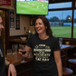 Woman in a bar wearing a humorous t-shirt, watching sports on TV.  The lady is wearing a Cows Corner t-shirt with the funny football slogan on the front that reads 'Your First Touch Is a Crime Against Football'. Cows Corner gifts are perfect for sport-mad fans, these gifts work brilliantly for birthdays, new baby celebrations, Father’s Day, Mother’s Day, Christmas, anniversaries, thank you gifts, end-of-season team awards, graduations, retirements, and just-because moments when you want to raise a smile.