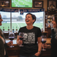 Three women in a bar watching sports on TV, one wearing a humorous t-shirt.  The lady is wearing a Cows Corner T-shirt with a funny football slogan on the front that reads 'Your Tekkers Are As Rare as a Unicorn Sighting'.  Cows Corner gifts are perfect for sport-mad fans, these gifts work brilliantly for birthdays, new baby celebrations, Father’s Day, Mother’s Day, Christmas, anniversaries, thank you gifts, end-of-season team awards, graduations, retirements, and just-because moments 