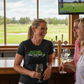 Woman holding a glass of wine in a bar with a view of a golf course on a TV screen. The lady is wearing a Cows Corner t-shirt with the funny golf saying on the front that reads 'Golf’s There Ugliest Words: ’Still Your Shot! – Dave Marr'.  Cows Corner gifts are perfect for sport-mad fans, these gifts work brilliantly for birthdays, new baby celebrations, Father’s Day, Mother’s Day, Christmas, anniversaries, thank you gifts, end-of-season team awards, graduations, retirements, and just-because moments 