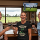 Three people in a bar setting, enjoying wine and watching a golf game on TV. The lady is wearing a Cows Corner t-shirt with a funny golf slogan 'Hole-In-One Achieved… Retirement Pending'. Cows Corner gifts are perfect for sport-mad fans, these gifts work brilliantly for birthdays, new baby celebrations, Father’s Day, Mother’s Day, Christmas, anniversaries, thank you gifts, end-of-season team awards, graduations, retirements, and just-because moments when you want to raise a smile. 
