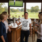 Woman in a bar wearing a t-shirt with humorous text, surrounded by friends with drinks, watching golf on TV. The lady is wearing a Cows Corner t-shirt with the funny golf slogan 'Hole-In-One Today… Pub Stories Forever'. Cows Corner gifts are perfect for sport-mad fans, these gifts work brilliantly for birthdays, new baby celebrations, Father’s Day, Mother’s Day, Christmas, anniversaries, thank you gifts, end-of-season team awards, graduations, retirements, and just-because moments 