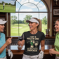 Three women in golf attire with a humorous t-shirt, standing in a room with a large window showing a golf course. The lady is wearing a Cows Corner t-shirt with a funny golf slogan 'I Got A Hole-In-One… My Ball’s Still Shocked'. Cows Corner gifts are perfect for sport-mad fans, these gifts work brilliantly for birthdays, new baby celebrations, Father’s Day, Mother’s Day, Christmas, anniversaries, thank you gifts, end-of-season team awards, graduations, retirements, and just-because moments 