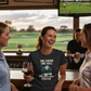 Three women in a bar with a TV showing a golf game, one wearing a humorous t-shirt. The lady is wearing a Cows Corner t-shirt with a funny golf slogan 'One Swing, One Hole, One Very Smug Golfer'. Cows Corner gifts are perfect for sport-mad fans, these gifts work brilliantly for birthdays, new baby celebrations, Father’s Day, Mother’s Day, Christmas, anniversaries, thank you gifts, end-of-season team awards, graduations, retirements, and just-because moments when you want to raise a smile. 
