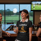 Woman in a golf-themed shirt laughing with friends in a bar with a golf course view. The lady is wearing a Cows Corner t-shirt with a funny golf saying on the front that reads 'A Bad Day at Golf Is Better Than a Good Day at Work'.  Cows Corner gifts are perfect for sport-mad fans, these gifts work brilliantly for birthdays, new baby celebrations, Father’s Day, Mother’s Day, Christmas, anniversaries, thank you gifts, end-of-season team awards, graduations, retirements, and just-because moments 