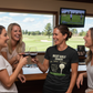 Four women in a room with a golf simulator, drinking wine and smiling. The lady is wearing a Cows Corner t-shirt with the golf coach slogan on the front that reads 'Best Golf Coach in the World. Thanks for Every Swing, Smile and Slice Fix'.  Cows Corner gifts are perfect for sport-mad fans, these gifts work brilliantly for birthdays, new baby celebrations, Father’s Day, Mother’s Day, Christmas, anniversaries, thank you gifts, end-of-season team awards, graduations, retirements, and just-because moments