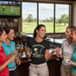 Group of women in a bar with golf-themed decor, watching a golf game on TV. The lady is wearing a Cows Corner T-shirt with the funny golf slogan on the front that reads 'I Like Big Putts and I Cannot Lie, but Yours Are Embarrassing'. Cows Corner gifts are perfect for sport-mad fans, these gifts work brilliantly for birthdays, new baby celebrations, Father’s Day, Mother’s Day, Christmas, anniversaries, thank you gifts, end-of-season team awards, graduations, retirements, and just-because moments
