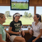 Three women sitting together in a golf-themed setting with a TV screen displaying a golf game. The lady is wearing a Cows Corner t-shirt with the funny golf slogan on the front that reads 'If Golf Was Easy You’d Be Good at It'. Cows Corner gifts are perfect for sport-mad fans, these gifts work brilliantly for birthdays, new baby celebrations, Father’s Day, Mother’s Day, Christmas, anniversaries, thank you gifts, end-of-season team awards, graduations, retirements, and just-because moments 