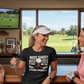 Two women in a golf-themed bar, one wearing a black t-shirt with a humorous golf-themed design.  The lady is wearing a Cows Corner t-shirt with the funny golf slogan on the front that reads 'Nice Shot! Too Bad It Was on the Wrong Fairway'. Cows Corner gifts are perfect for sport-mad fans, these gifts work brilliantly for birthdays, new baby celebrations, Father’s Day, Mother’s Day, Christmas, anniversaries, thank you gifts, end-of-season team awards, graduations, retirements, and just-because moments 
