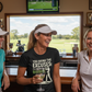Three women in a casual setting with a TV screen showing a golf game in the background. The lady is wearing a Cows Corner t-shirt with the funny golf slogan on the fron that reads 'You Bring the Excuses, I’ll Bring the Birdies'. Cows Corner gifts are perfect for sport-mad fans, these gifts work brilliantly for birthdays, new baby celebrations, Father’s Day, Mother’s Day, Christmas, anniversaries, thank you gifts, end-of-season team awards, graduations, retirements, and just-because moments 