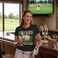 Woman in a golf-themed t-shirt holding a glass of wine in a bar with a TV showing a golf game.  The lady is wearing a Cows Corner t-shirt with the funny golf slogan on the front that reads 'You’d Be Great at Golf if It Wasn’t for the Hitting Part'.  Cows Corner gifts are perfect for sport-mad fans, these gifts work brilliantly for birthdays, new baby celebrations, Father’s Day, Mother’s Day, Christmas, anniversaries, thank you gifts, end-of-season team awards, graduations, retirements, and just-because