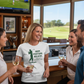 Group of people in a bar watching golf on TV with a woman wearing a Cows Corner t-shirt with the golf coach slogan on the front that reads 'Grip, Posture, Believe. Thank You for Everything Coach'. Cows Corner gifts are perfect for sport-mad fans, these gifts work brilliantly for birthdays, new baby celebrations, Father’s Day, Mother’s Day, Christmas, anniversaries, thank you gifts, end-of-season team awards, graduations, retirements, and just-because moments when you want to raise a smile. 