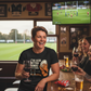People enjoying drinks and watching a sports match in a pub. The lady is wearing a Cows Corner t-shirt with the funny rugby slogan 'I’ve Seen Better Hands on a Clock – Phil Kearns'. Cows Corner gifts are perfect for sport-mad fans, these gifts work brilliantly for birthdays, new baby celebrations, Father’s Day, Mother’s Day, Christmas, anniversaries, thank you gifts, end-of-season team awards, graduations, retirements, and just-because moments when you want to raise a smile. 

