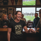 Woman wearing a humorous t-shirt in a pub with people and a TV in the background, the lady is wearing a Cows Corner t-shirt with the funny rugby slogan 'A Rugby Tour Is Like Sex. When It’s Good, It’s Great. When It’s Bad, It’s Still Pretty Good – Willie John McBride'. Cows Corner gifts are perfect for sport-mad fans, these gifts work brilliantly for birthdays, new baby celebrations, Father’s Day, Mother’s Day, Christmas, anniversaries, thank you gifts, end-of-season team awards, graduations, retirements