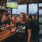 Woman in a bar wearing a black t-shirt with rugby-themed text, surrounded by people watching a game on TV. The lady is wearing a Cows Corner t-shirt with the funny rugby slogan on the front that reads 'If Rugby Was Easy They’d Call It Football'. Cows Corner gifts are perfect for sport-mad fans, these gifts work brilliantly for birthdays, new baby celebrations, Father’s Day, Mother’s Day, Christmas, anniversaries, thank you gifts, end-of-season team awards, graduations, retirements, and just-because moments 