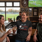 People in a pub watching sports on TV, with a woman in a black t-shirt with text. The lady is wearing a Cows Corner t-shirt with the funny rugby slogan on the front that reads 'No Guts No Glory No Teeth No Problem'. Cows Corner gifts are perfect for sport-mad fans, these gifts work brilliantly for birthdays, new baby celebrations, Father’s Day, Mother’s Day, Christmas, anniversaries, thank you gifts, end-of-season team awards, graduations, retirements, and just-because moments when you want to raise a smile