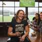 Woman in a rugby-themed t-shirt holding a glass of wine in a bar with a TV showing a sports game. The lady is wearing a Cows Corner t-shirt with the funny rugby slogan 'Rugby Is a Hooligan’s Game Played by Gentleman, and Football Is a Gentleman’s Game Played by Hooligans'. Cows Corner gifts are perfect for sport-mad fans, these gifts work brilliantly for birthdays, new baby celebrations, Father’s Day, Mother’s Day, Christmas, anniversaries, thank you gifts, end-of-season team awards, graduations, retirement